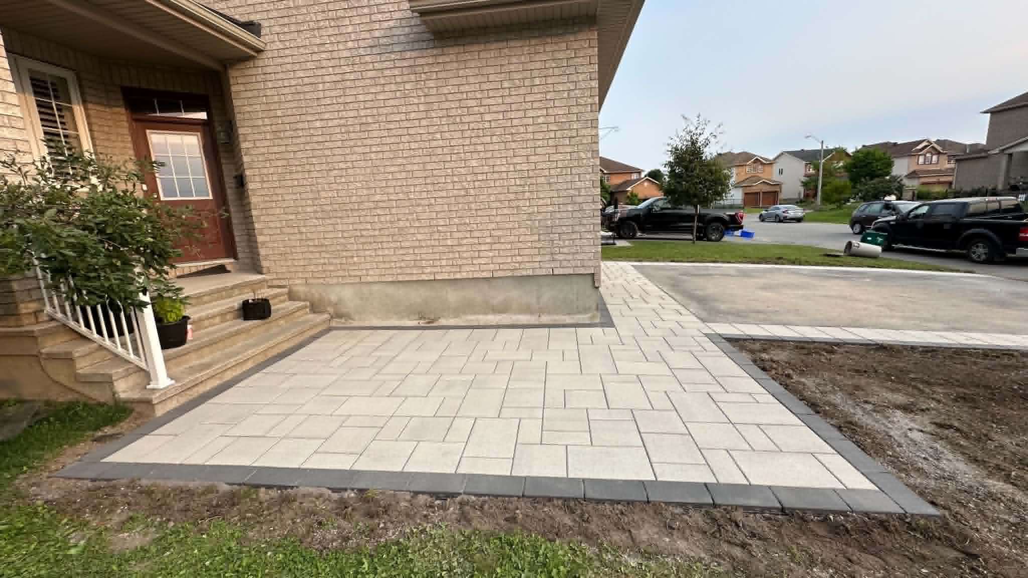 Light paver parking pad with a double charcoal border beside a brick house and driveway