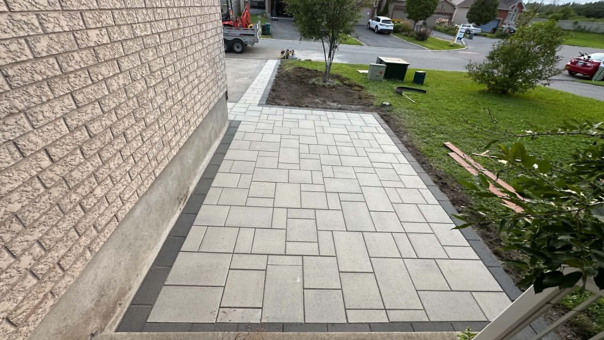 Beige paver extension pad with a charcoal border running from the driveway along the garage wall