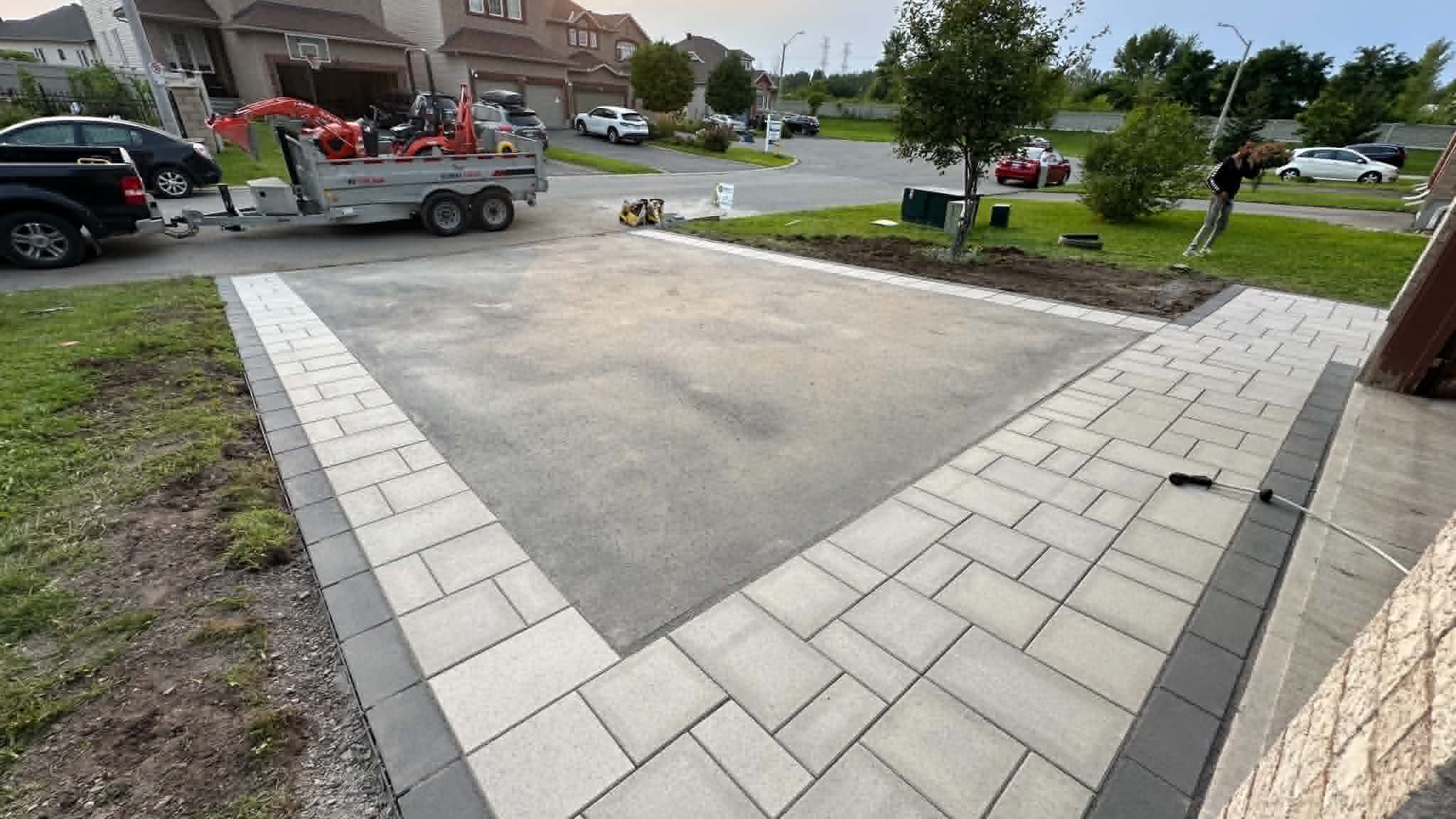 Driveway mid-install with paver borders laid around a compacted gravel base and a dump trailer at the street