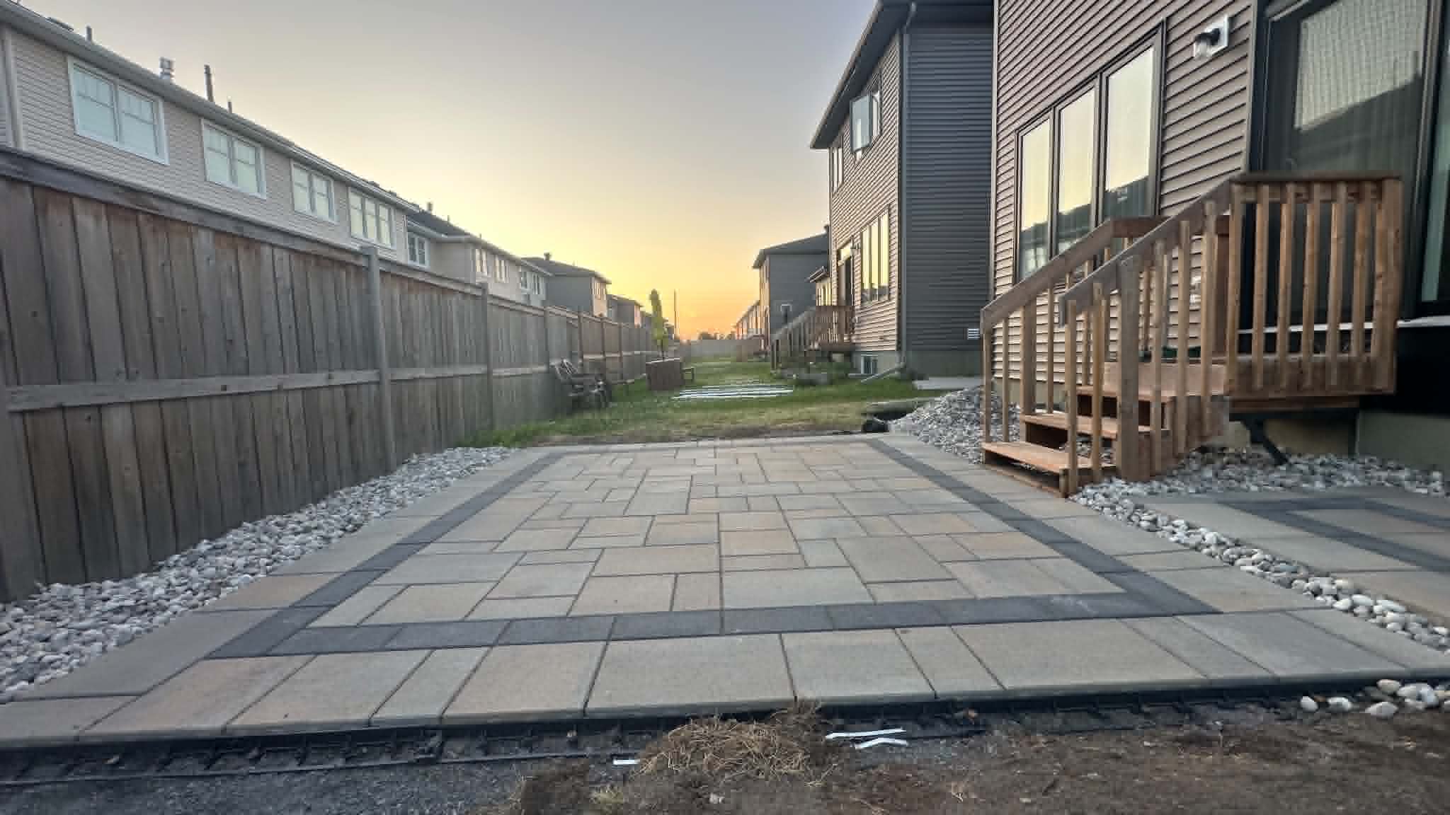 Backyard paver patio at dusk framed by a charcoal border course and river rock