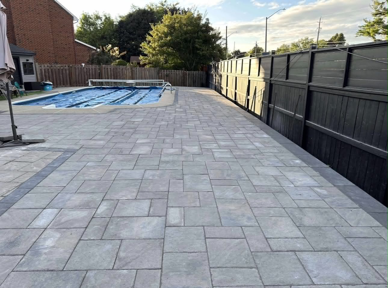 Grey paver pool deck wrapping around an inground pool beside a dark board fence