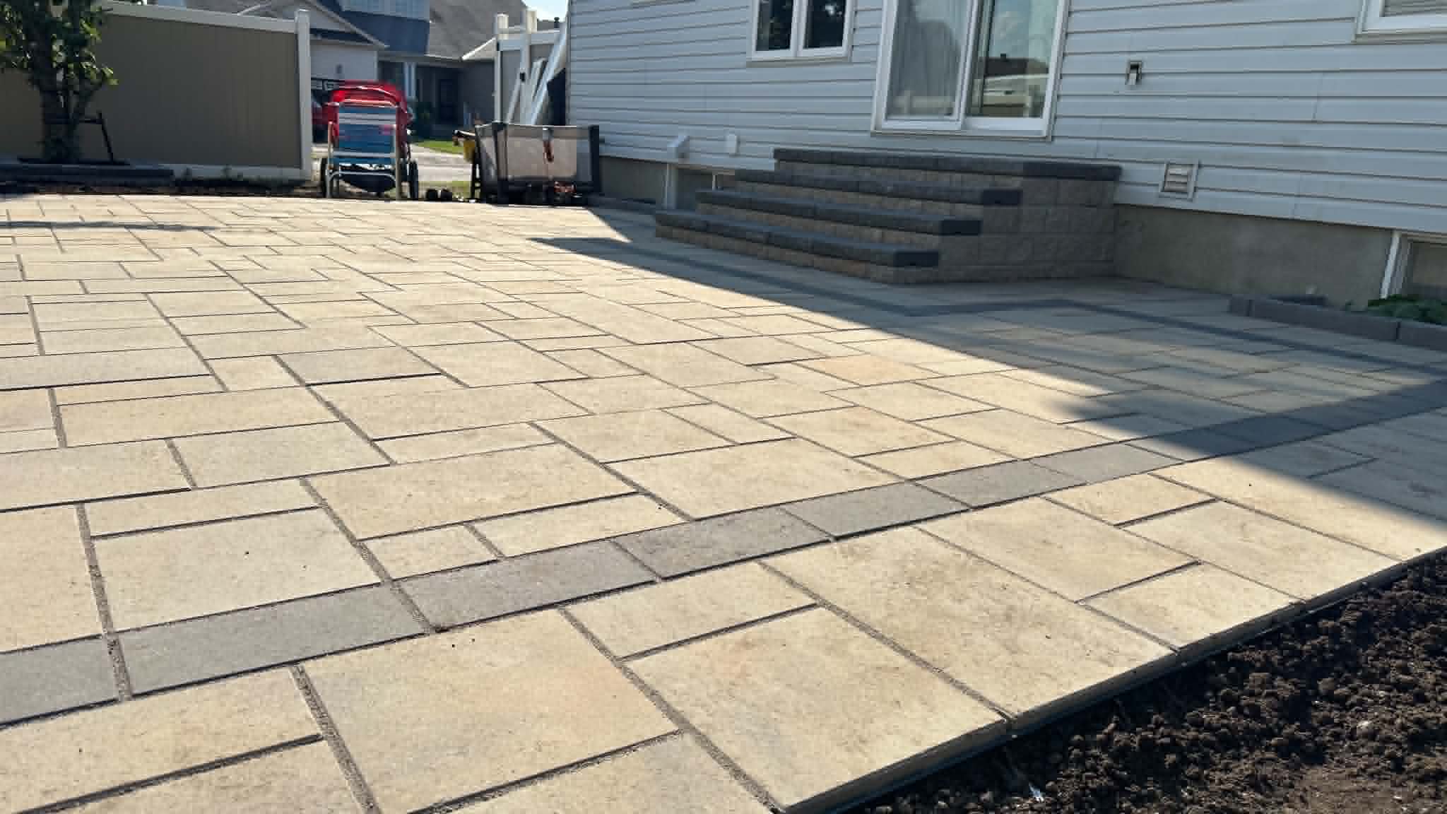 Wide beige slab patio with charcoal accent bands and paver steps up to a rear door