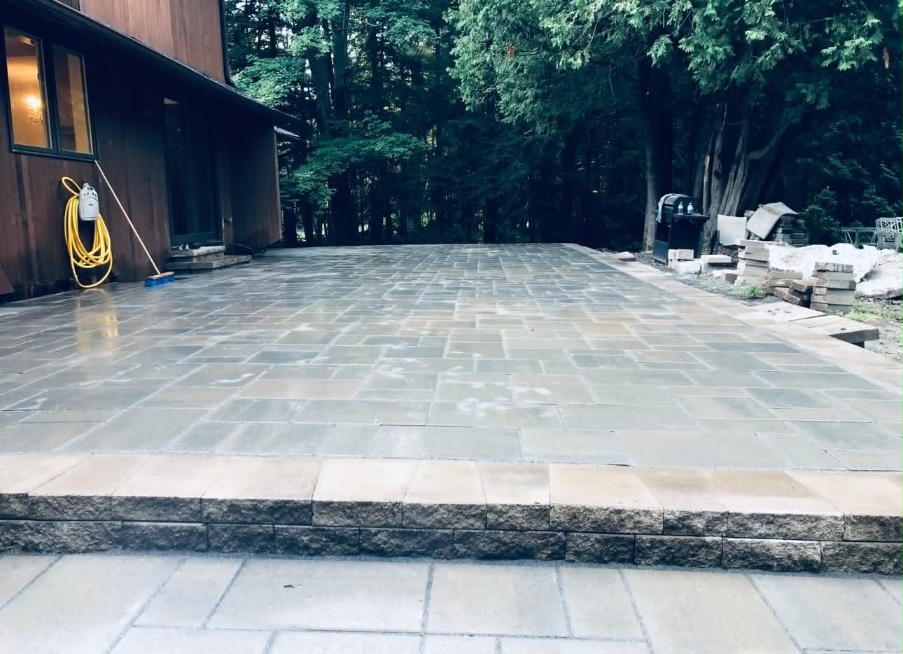 Wet grey patio in a wooded backyard stepped down over a rough-faced stone riser