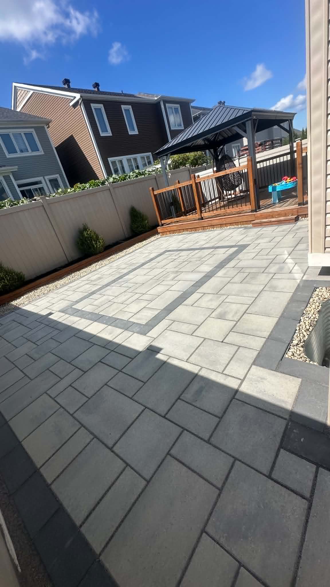 Grey paver patio with an inset border pattern beside a cedar deck and planter beds
