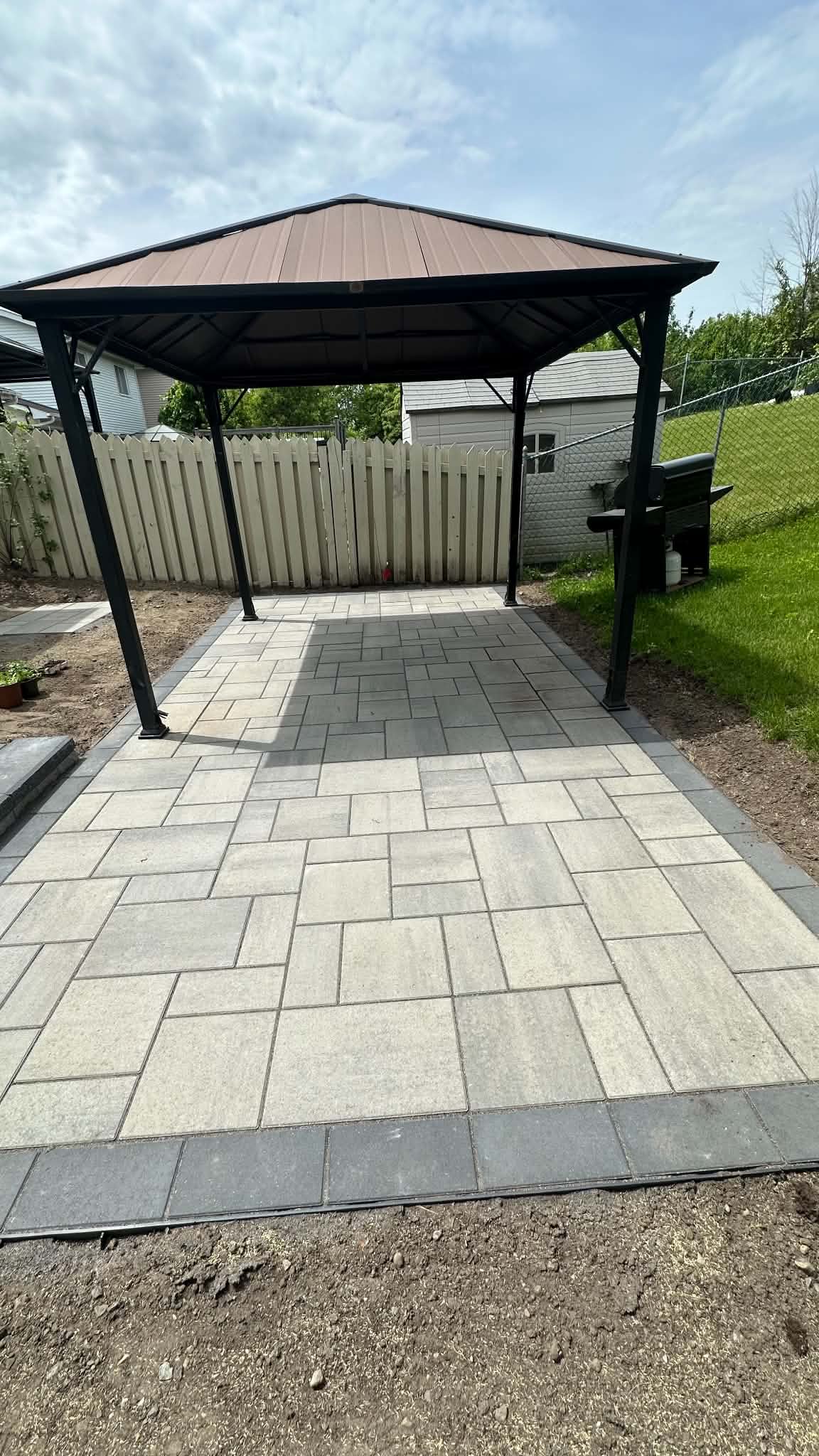Light grey paver patio under a black hard-top gazebo, finished with a charcoal border