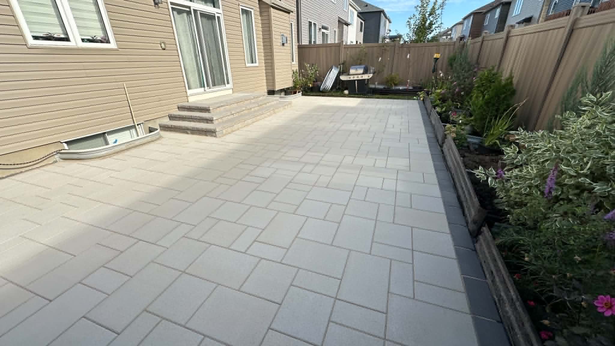 Long beige slab patio with a charcoal edge running past planter beds along a board fence