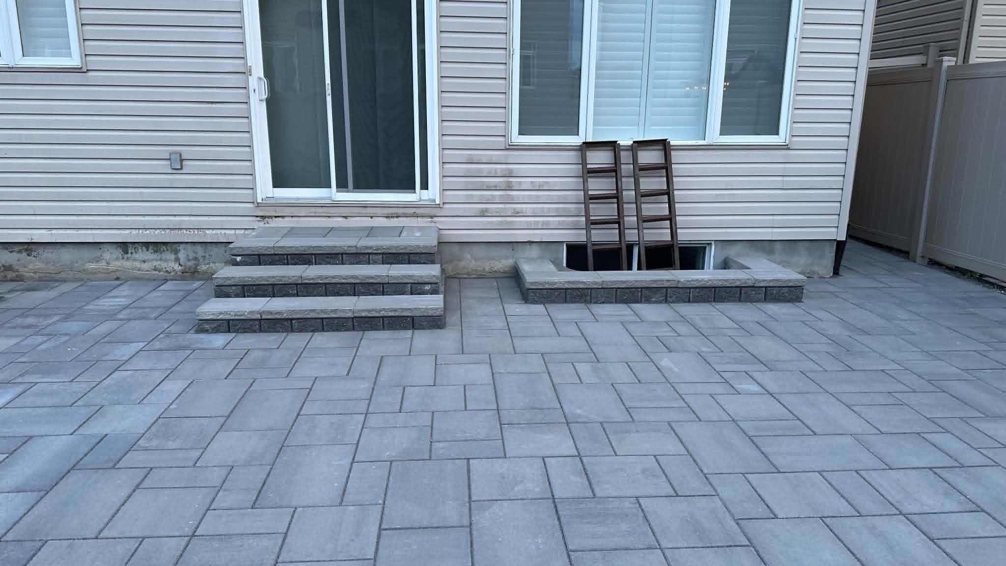 Grey random-pattern slab patio with paver steps at the sliding door and a low block wall around the window well