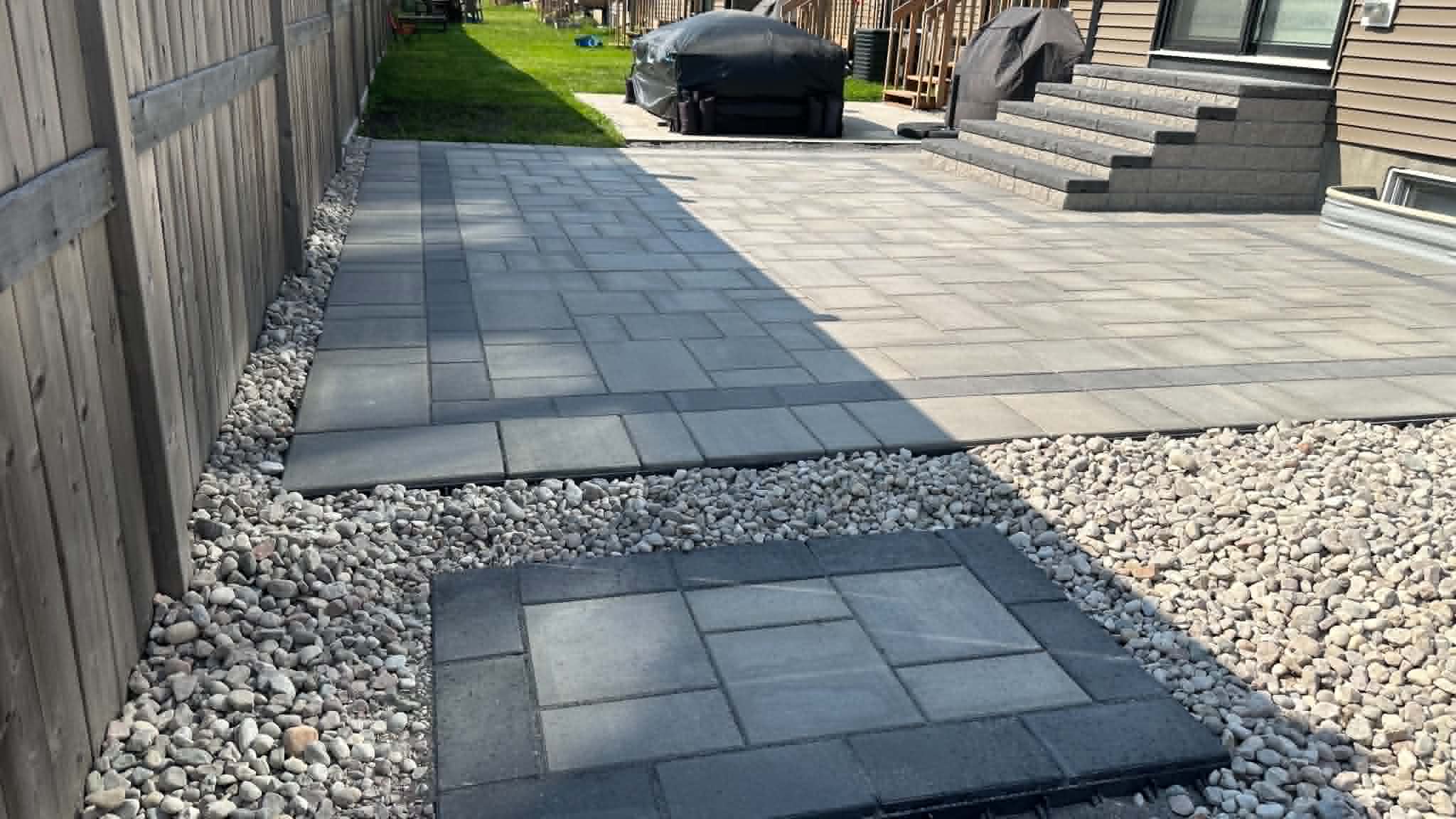 Two-tone backyard patio and a dark paver walk edged in river rock, with a covered hot tub and steps at the house