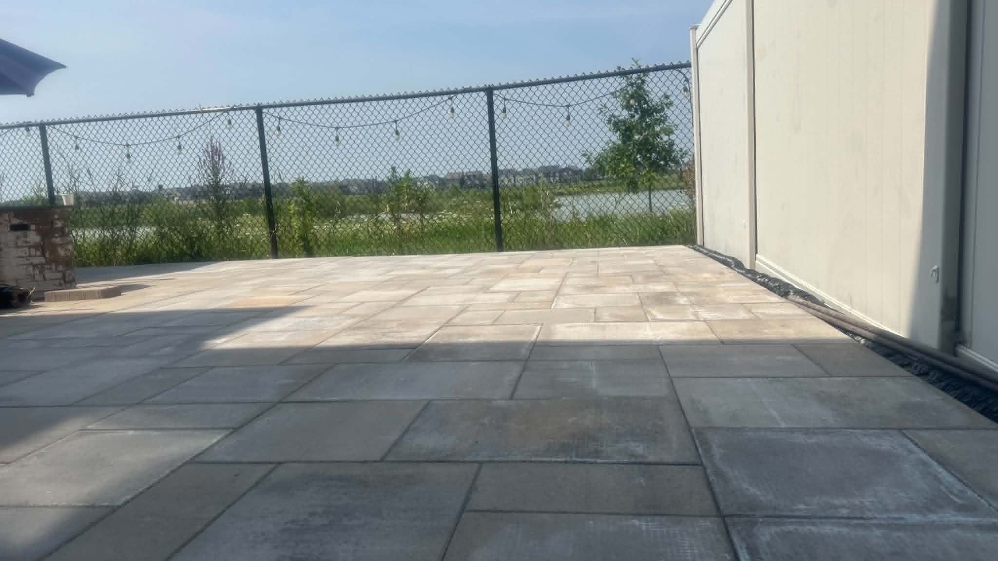 Charcoal and beige slab patio with string lights along a chain-link fence overlooking a pond