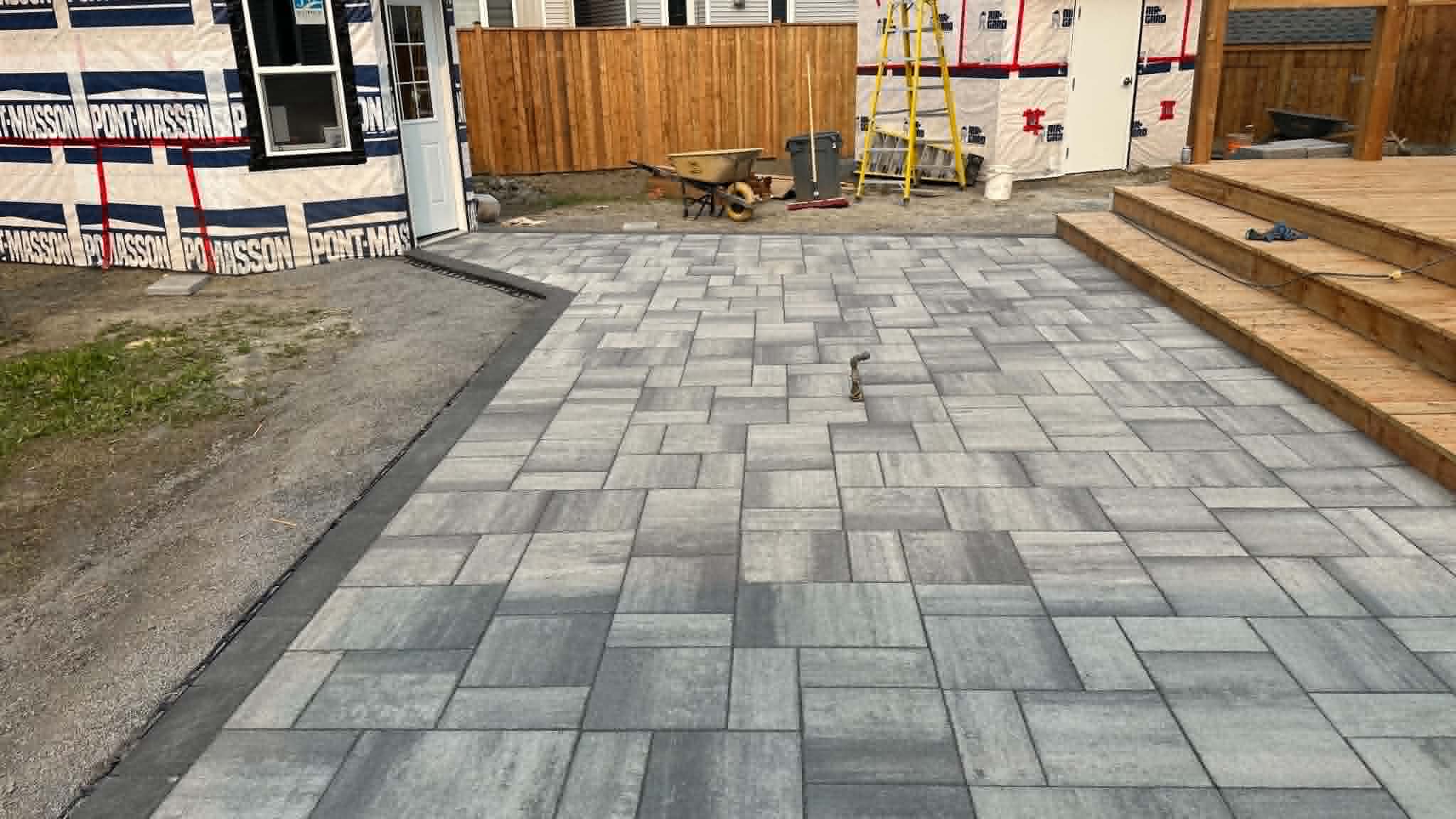 Grey two-tone paver patio at the wood deck steps of a house still in construction wrap
