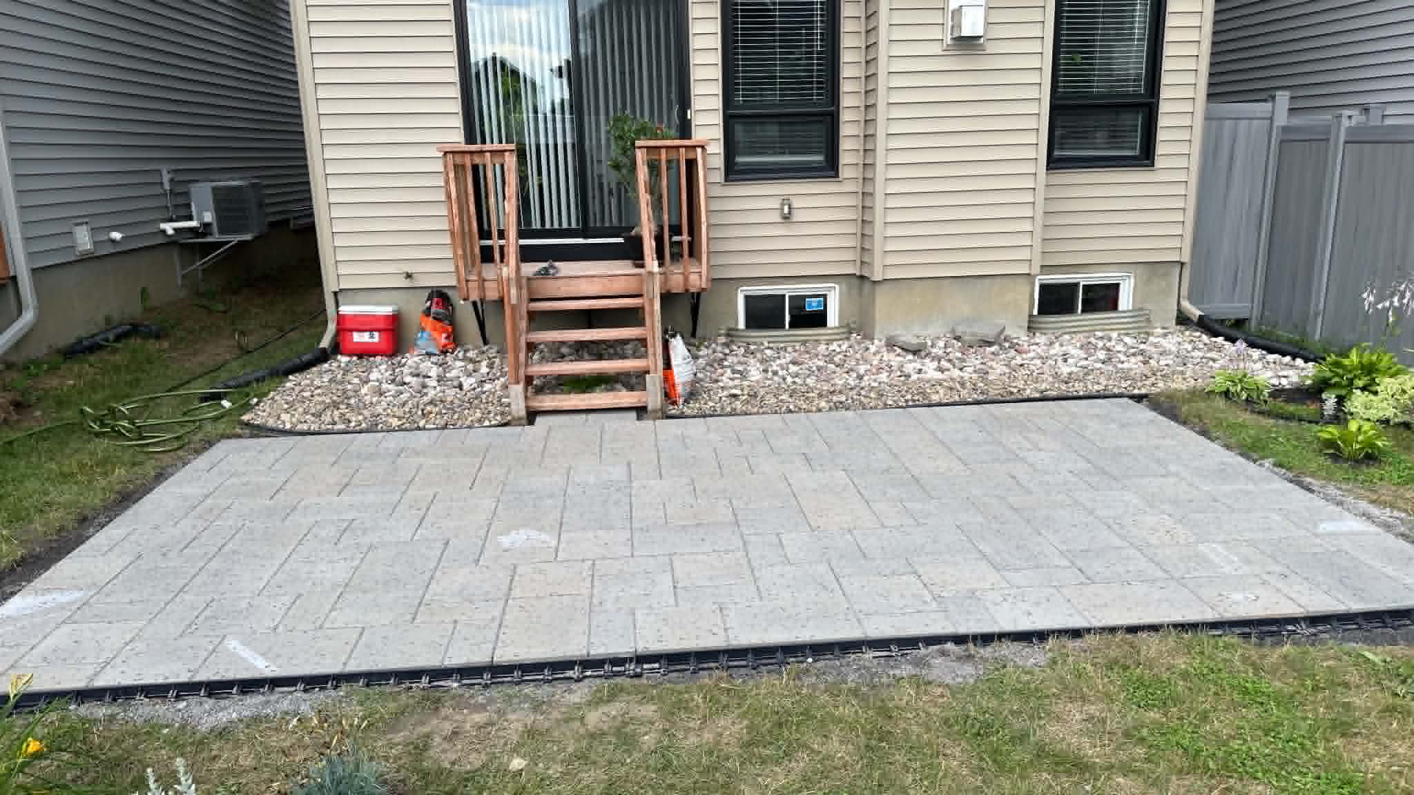 Grey paver patio pad at the base of wood deck stairs with river rock dressing the window wells