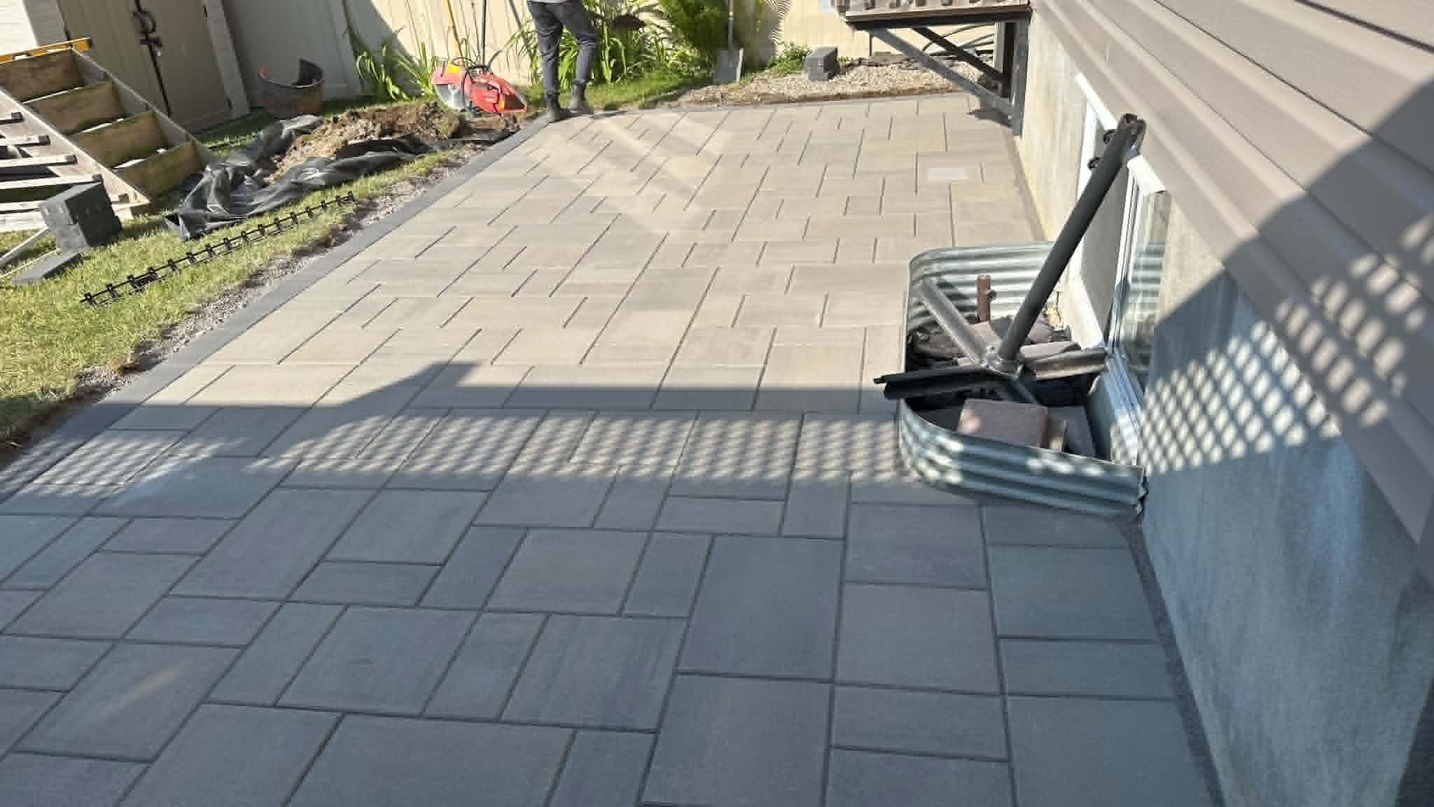 Grey and charcoal slab patio mid-install with edge restraint still exposed along the lawn