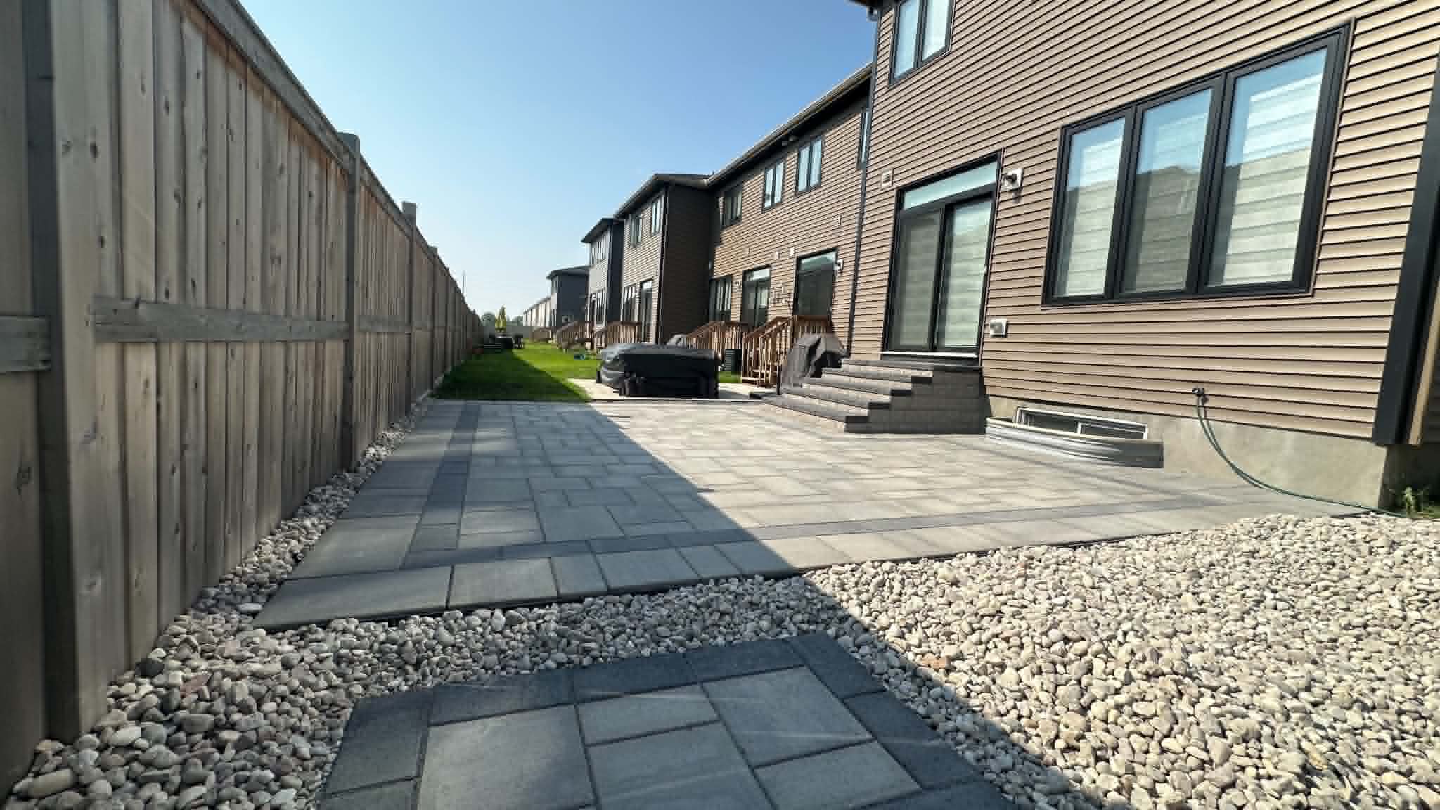 Long side-yard paver walkway and patio bordered by river rock beside a board fence