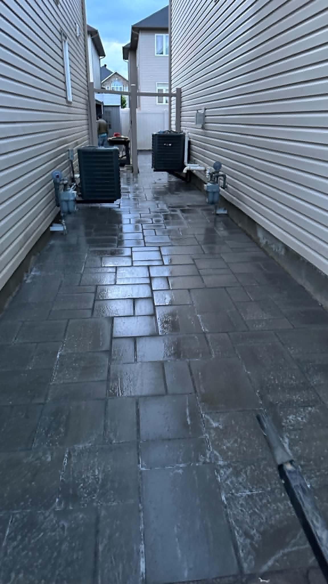 Wet dark paver walkway between two homes, freshly compacted with sand still on the surface
