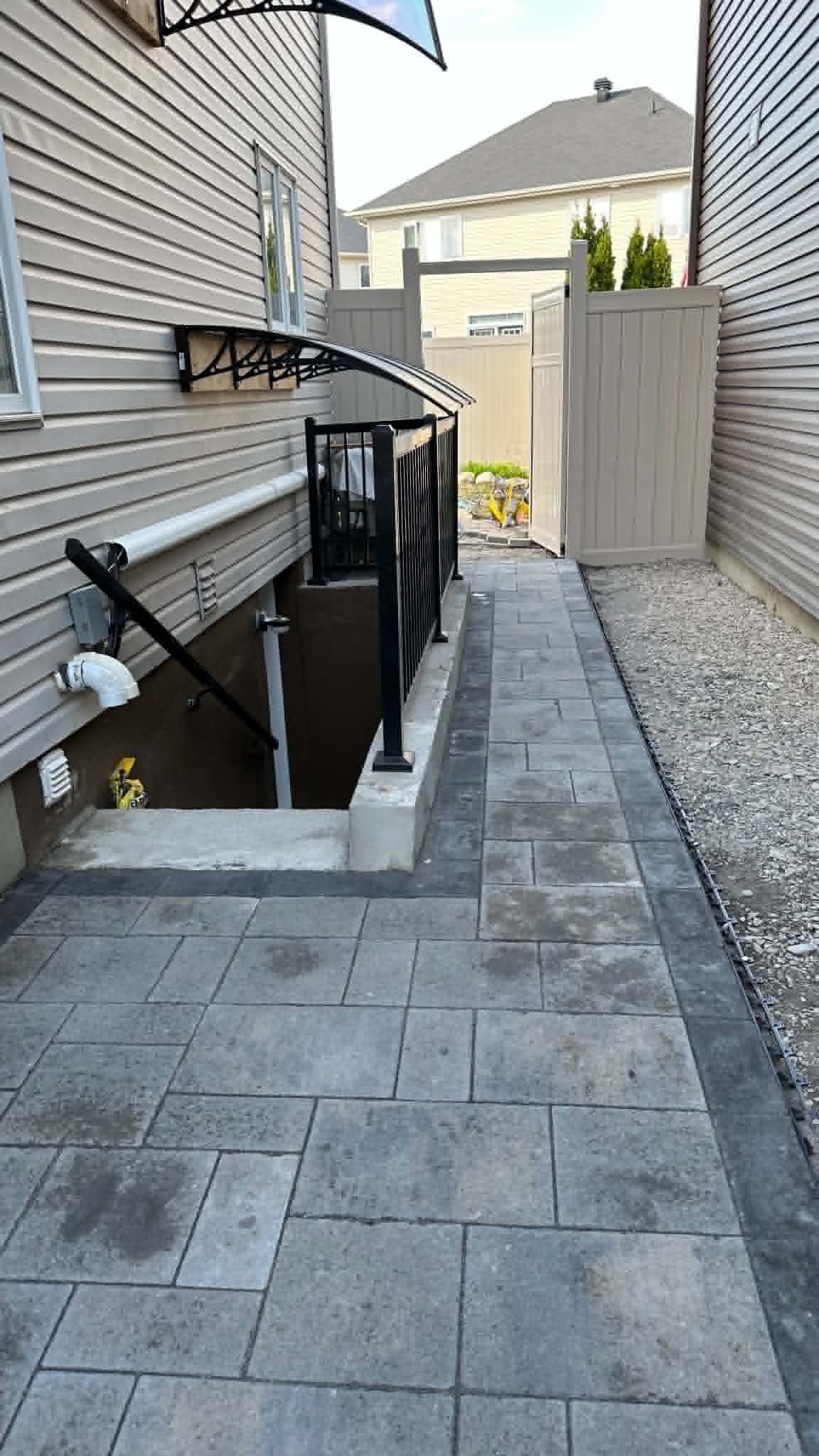 Bordered paver walk running past a railed basement stairwell to the backyard gate